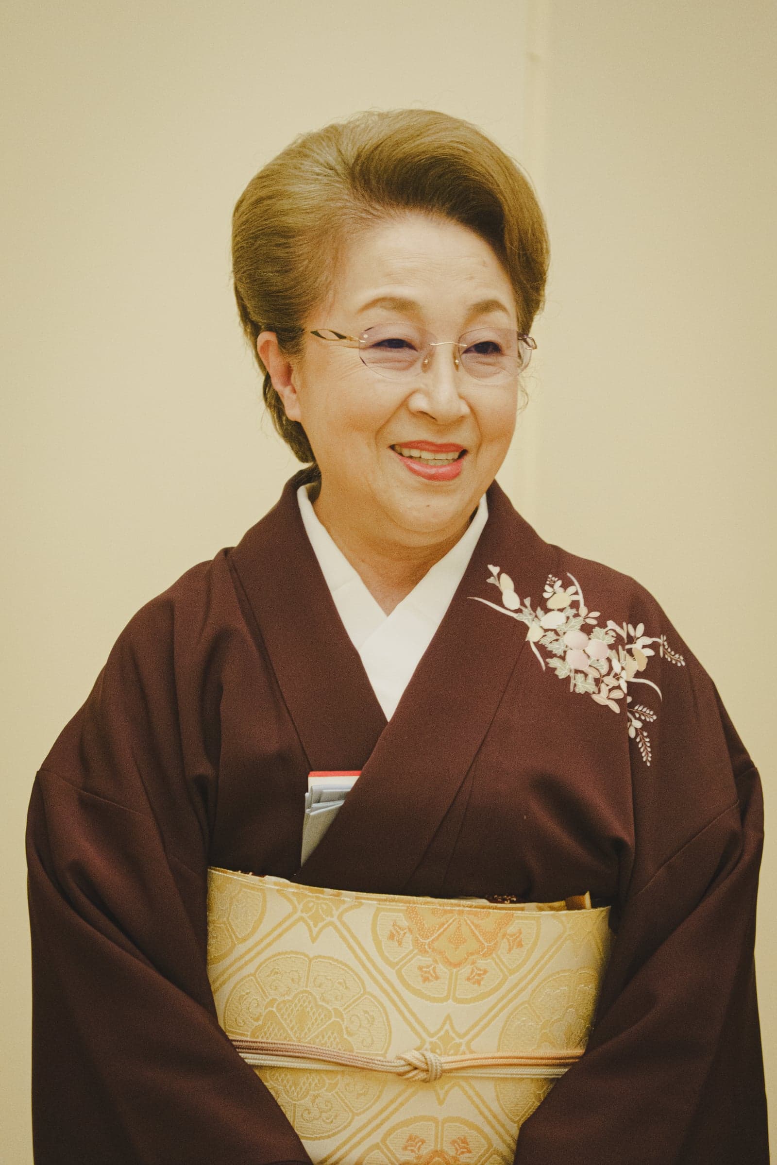 Okami Chikage smiling, in glasses and a deep red kimono with a gold obi.