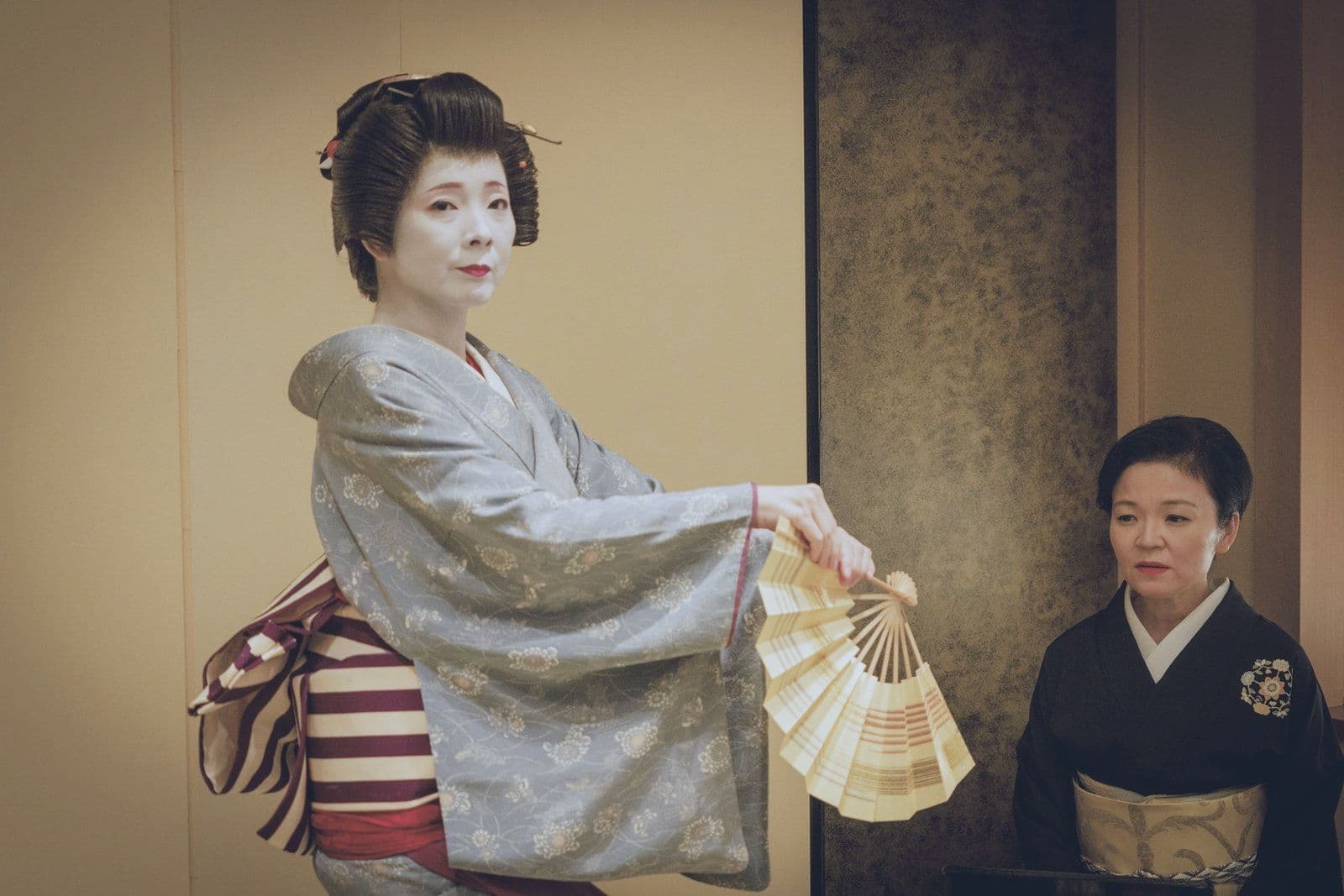 A geisha in a grey kimono dances with an open fan, a second geisha kneeling beside her.