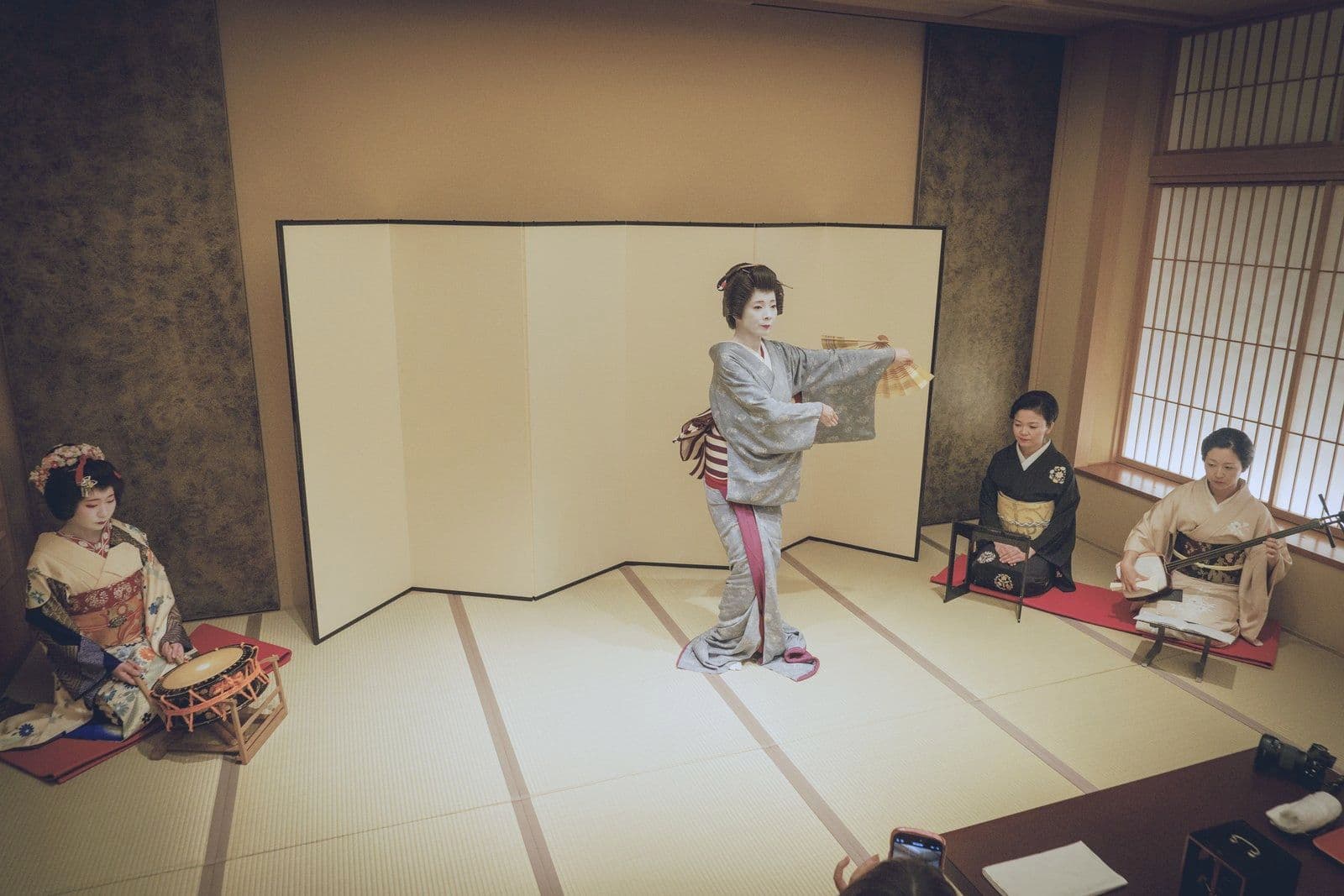 In a tatami room, a geisha dances before a gold folding screen while others wait with a drum and a shamisen.