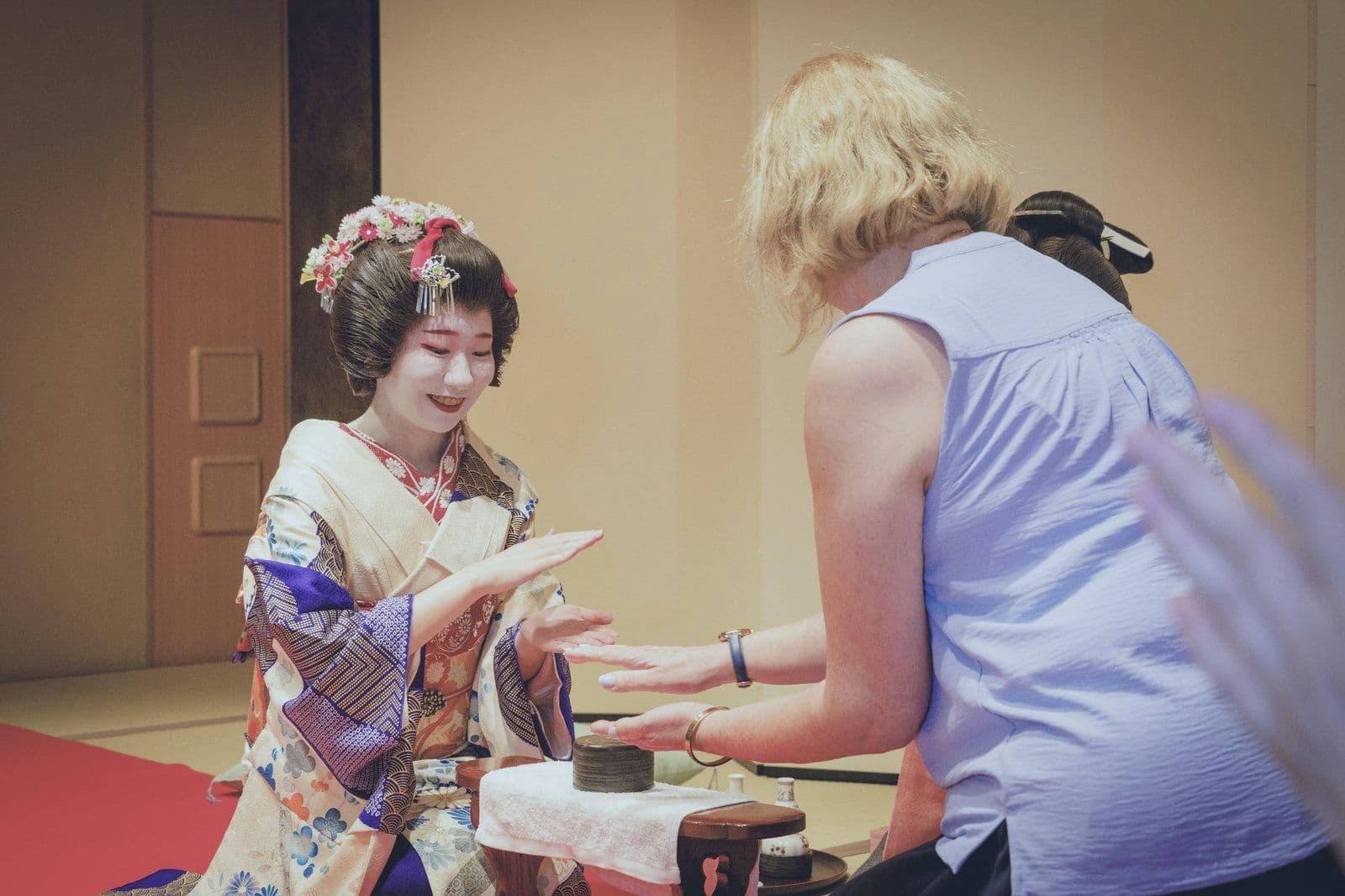A young geisha in a bright kimono plays konpira fune fune with a guest, seen from behind.