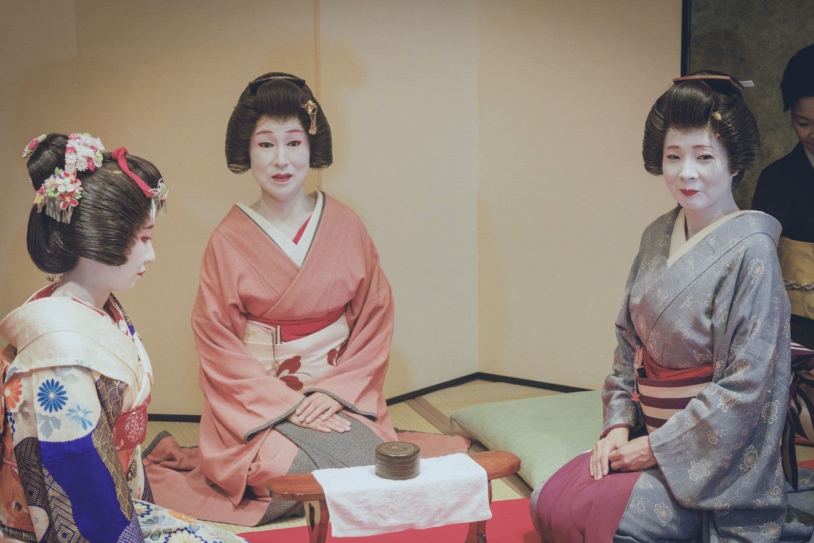 Three geisha in pale kimono sit side by side on the tatami, smiling for a photo.