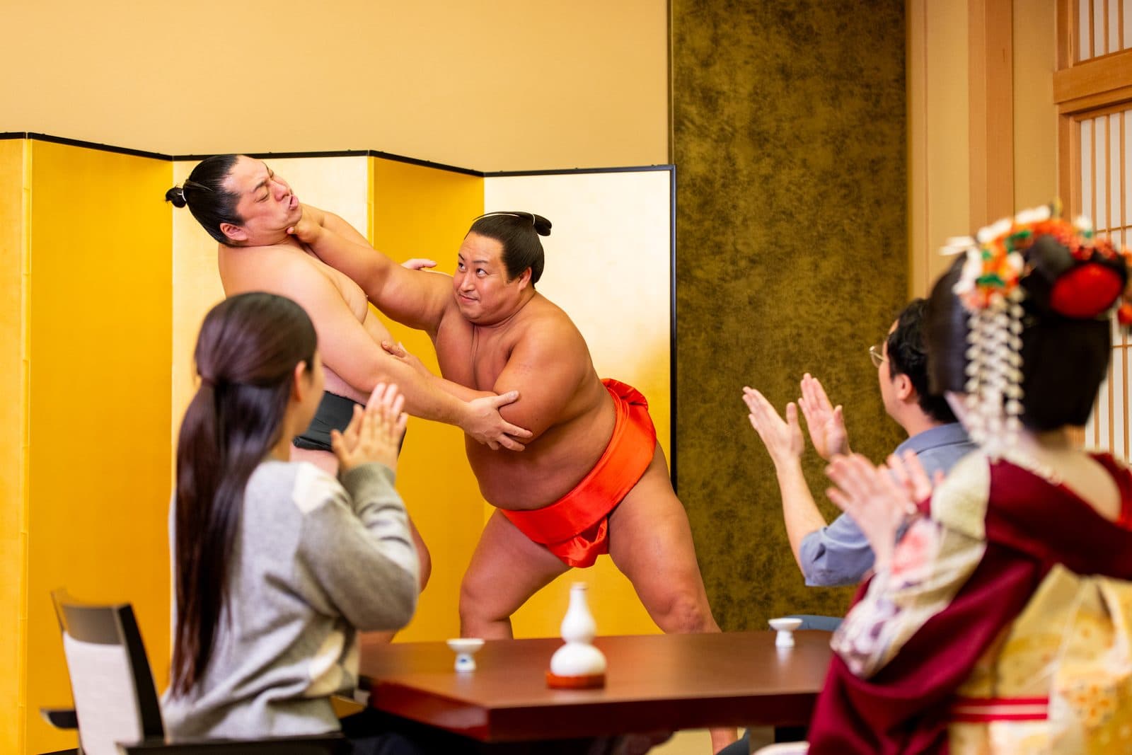 Two sumo actors demonstrate a bout beside the table as guests and a geisha applaud.