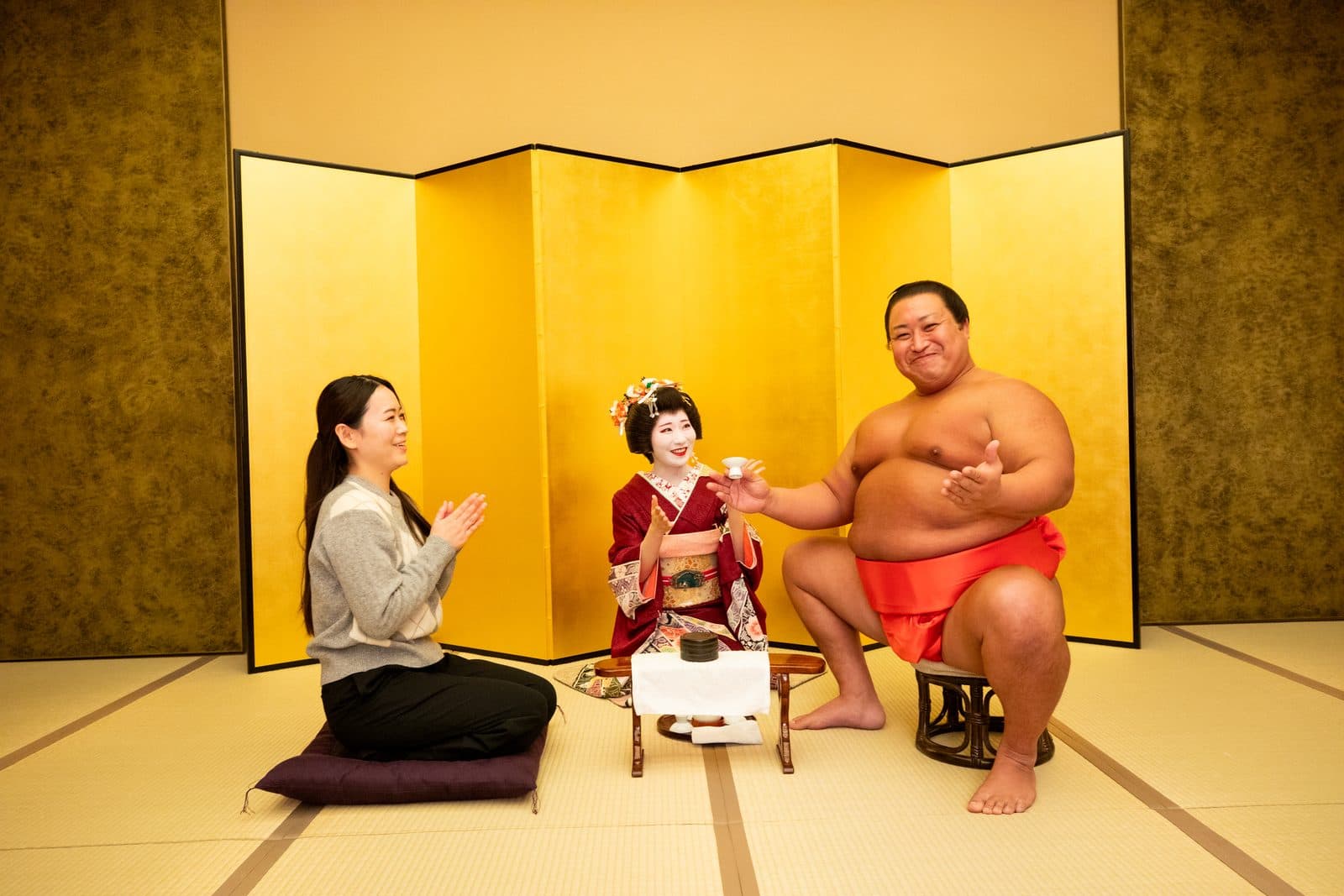 A guest and a geisha play an ozashiki game with a smiling sumo actor.