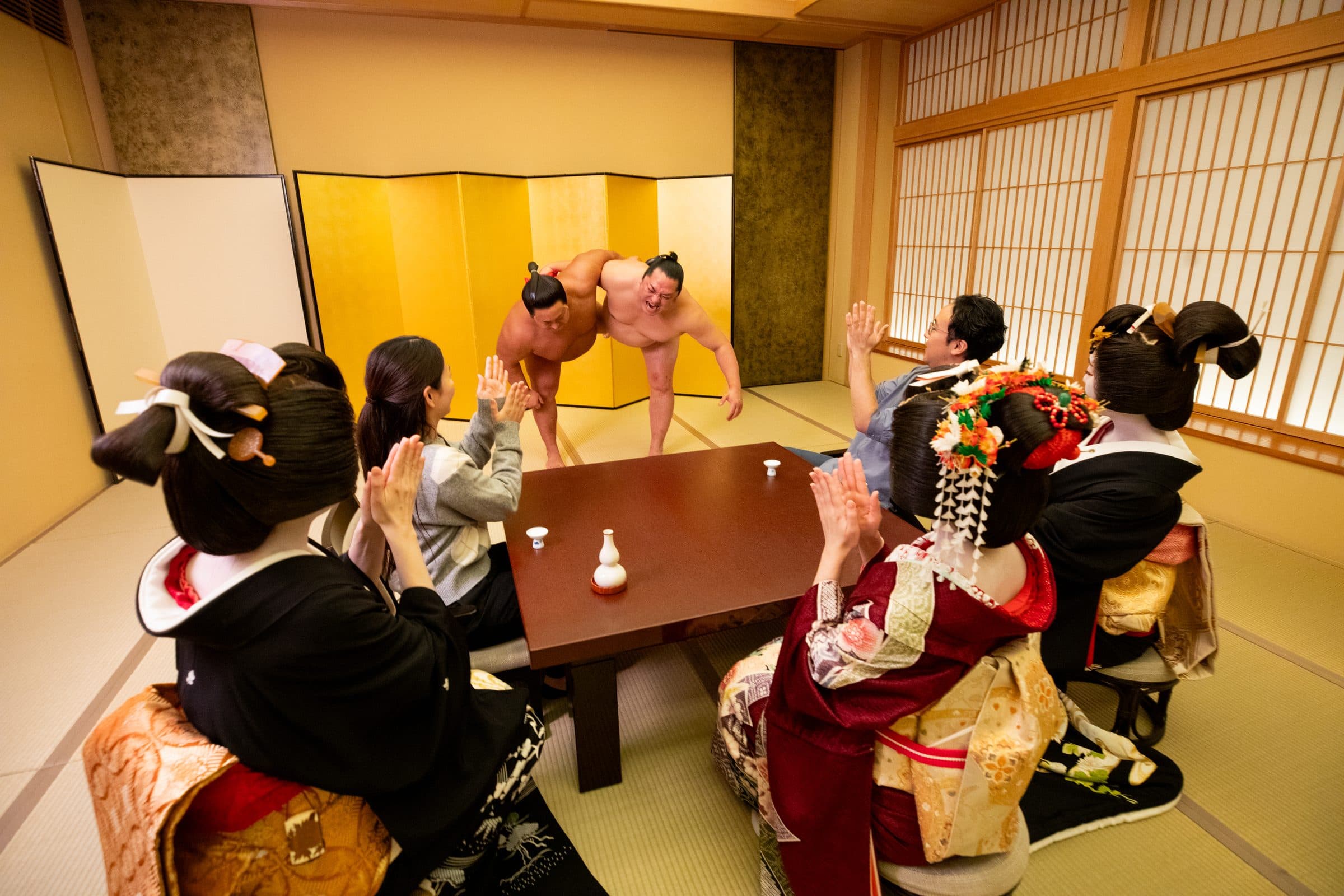 Sumo actors demonstrate a bout beside a low table, surrounded by guests and Asakusa geisha.