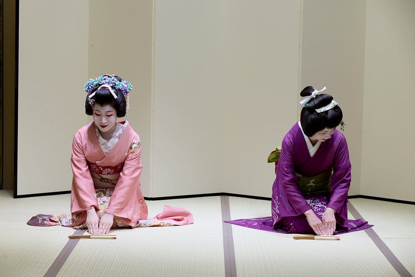 Two geisha kneel on the tatami and bow, their folded fans laid in front of them.