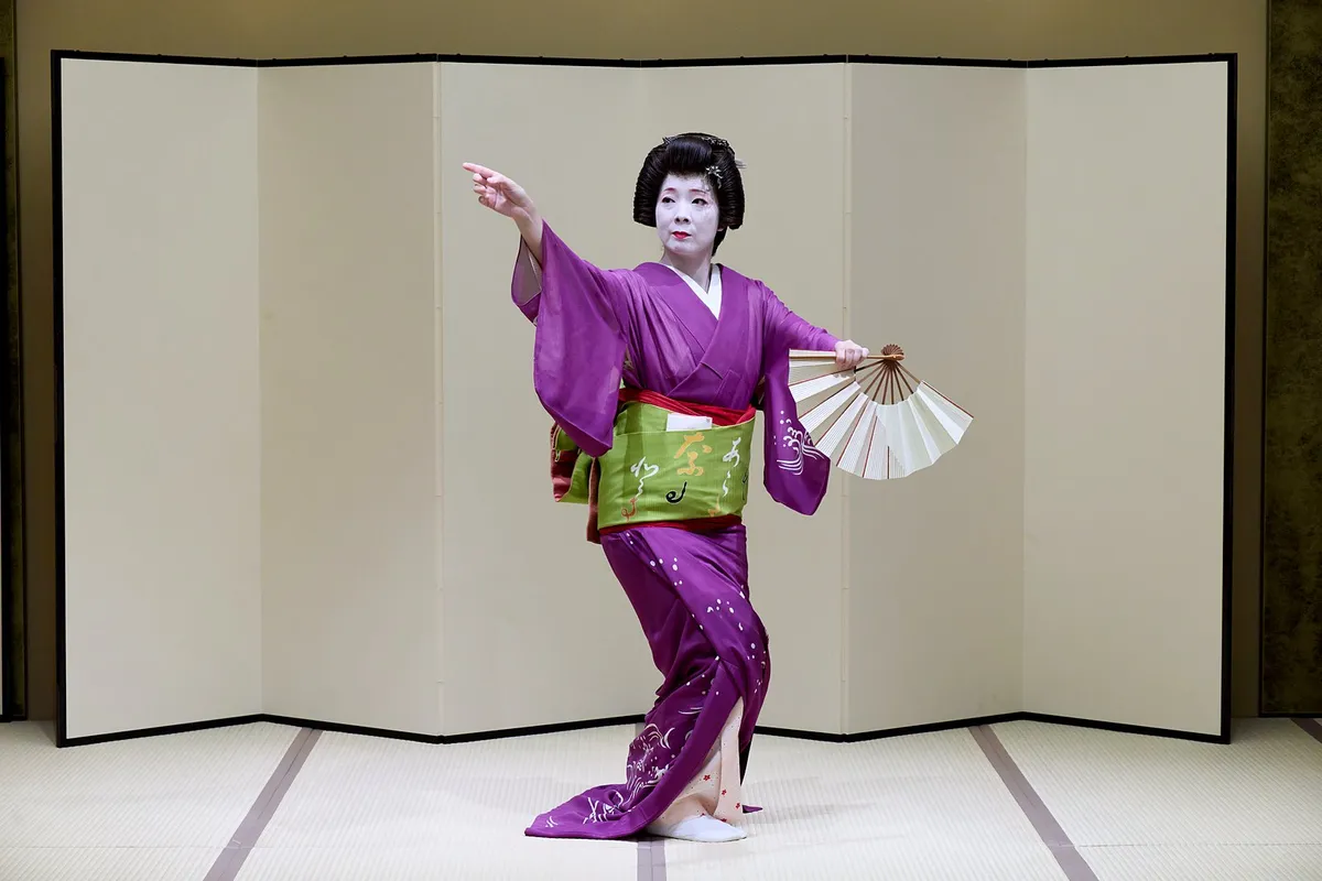 An Asakusa geisha dancing in front of a folding screen, one arm stretched out and an open fan in the other hand.