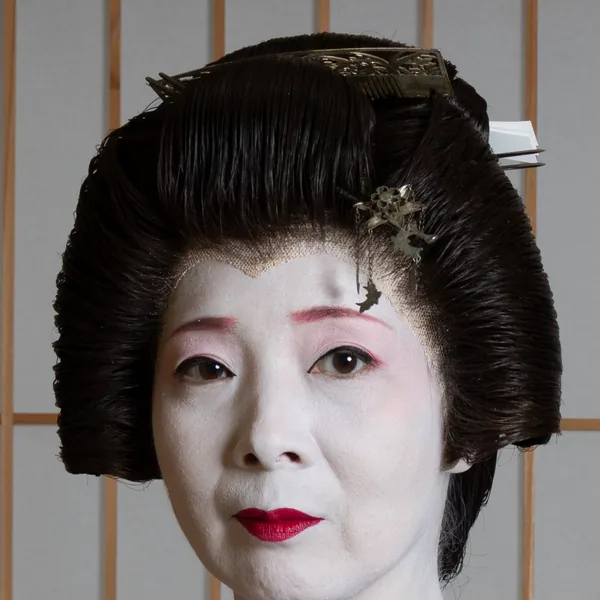 Close-up of a geisha's face in white makeup, red at the eyes and lips, with a comb and a hairpin in her nihongami.