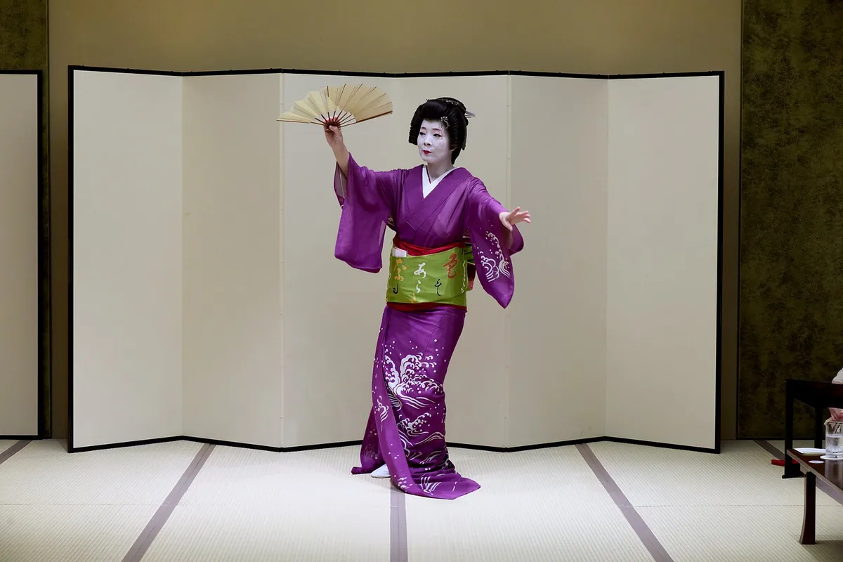 A tachikata dancing with a raised fan, the hem of her purple kimono trailing on the tatami.