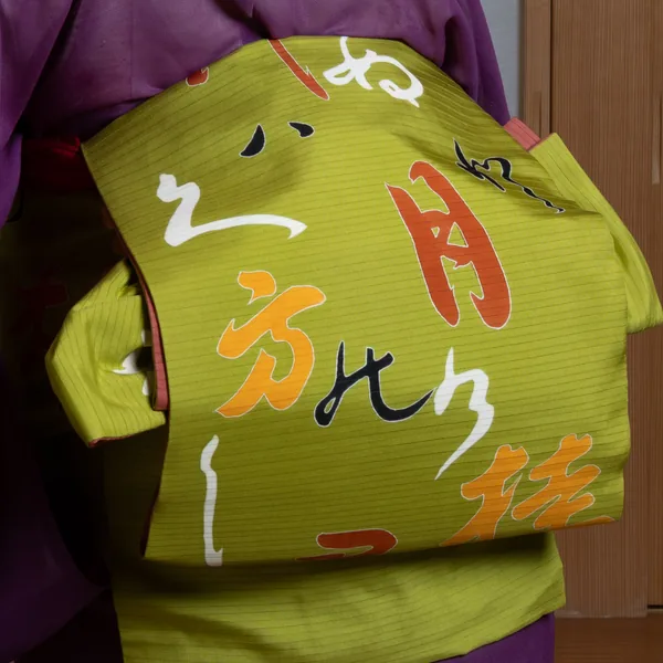 Close-up of a geisha from behind, a green obi with large written characters tied in an otaiko knot on the back of a purple kimono.