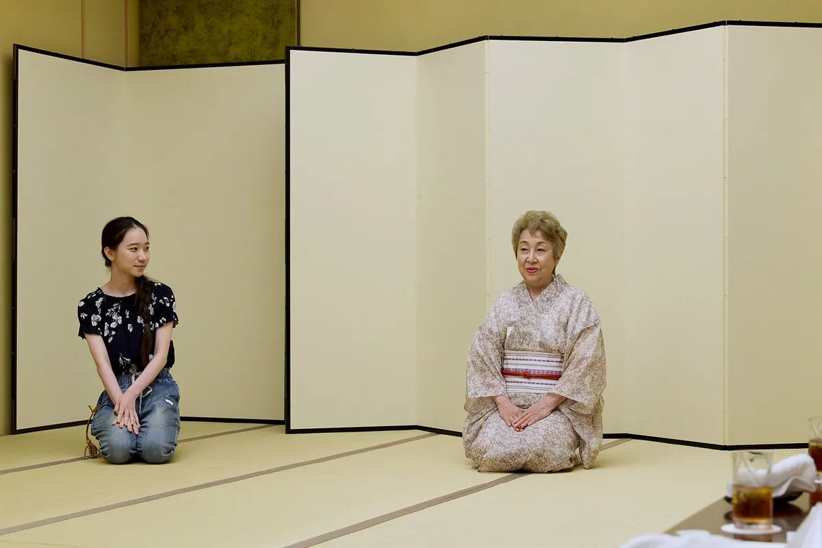 The okami seated on the tatami in front of a folding screen, talking, with a guest seated a little apart.