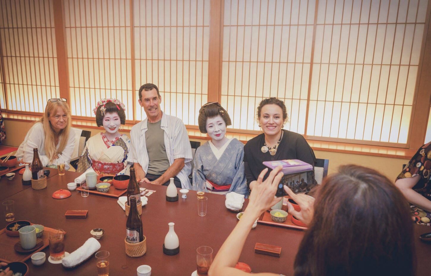 Group of people seated around a long dining table with ceramic cups and bottles; two women in traditional geisha makeup and kimono sit among smiling guests in a Japanese-style room.