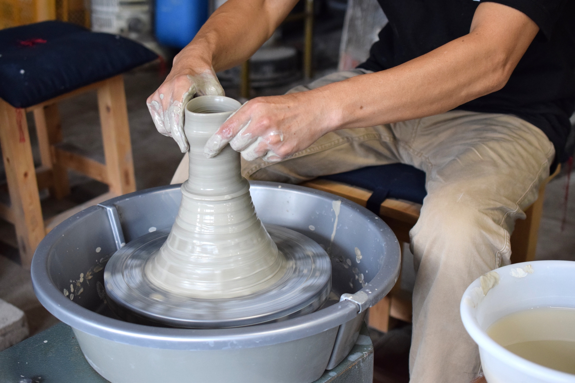 A potter's clay-covered hands shaping a vessel on a spinning pottery wheel in a workshop