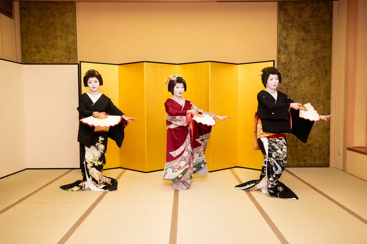 Three geisha-style performers in colorful kimonos perform a traditional dance on a tatami stage with a gold folding screen behind them.