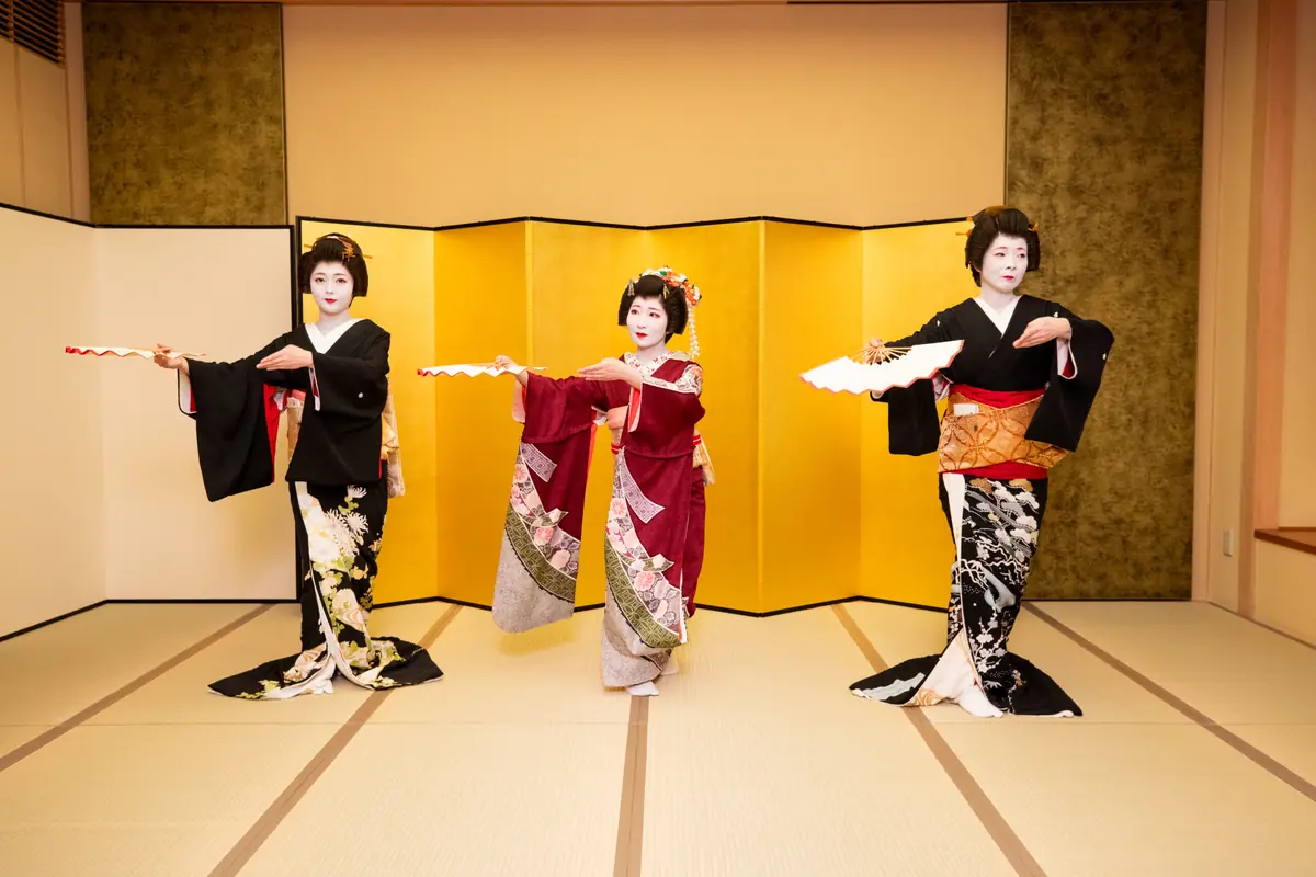 Three women in elegant kimonos perform a traditional fan dance in front of a gold folding screen.