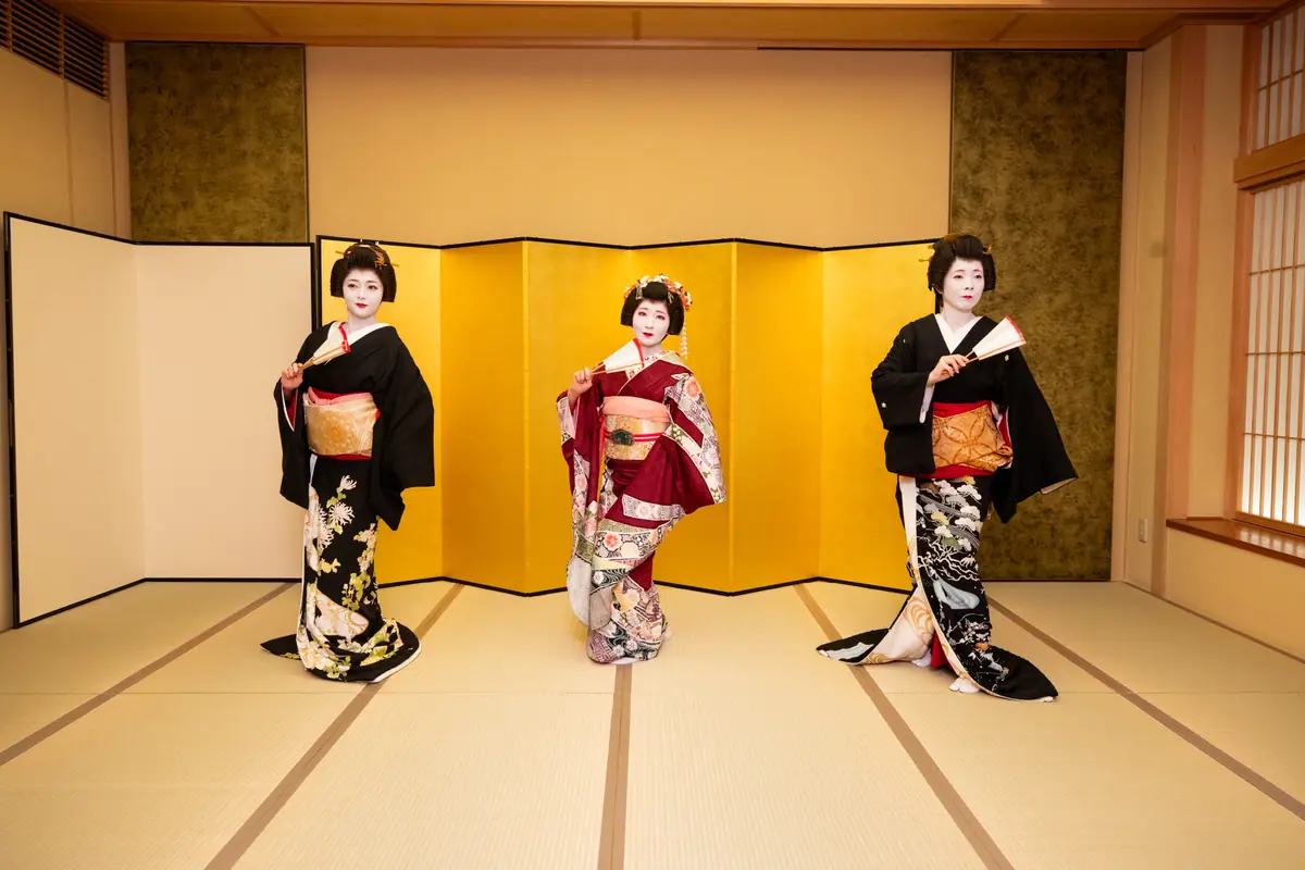 Three geishas in ornate kimonos pose on a tatami stage with a gold folding screen behind them, in a traditional room.