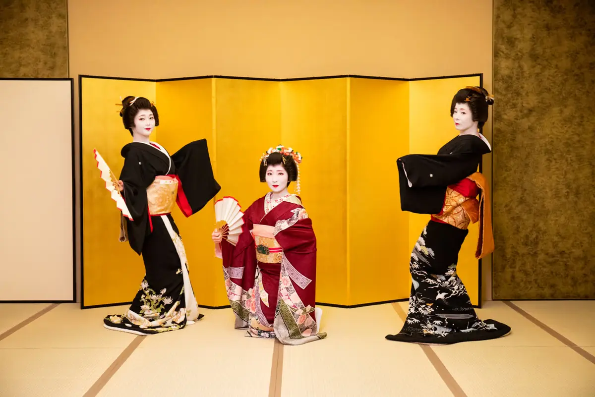 Three geisha in ornate kimonos pose with fans in front of a gold folding screen set.