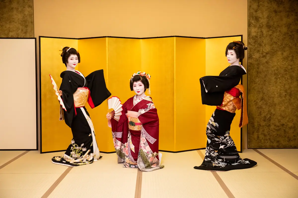 Three Geisha in elegant kimonos pose in front of a gold folding screen, with the center geisha kneeling and holding a fan.