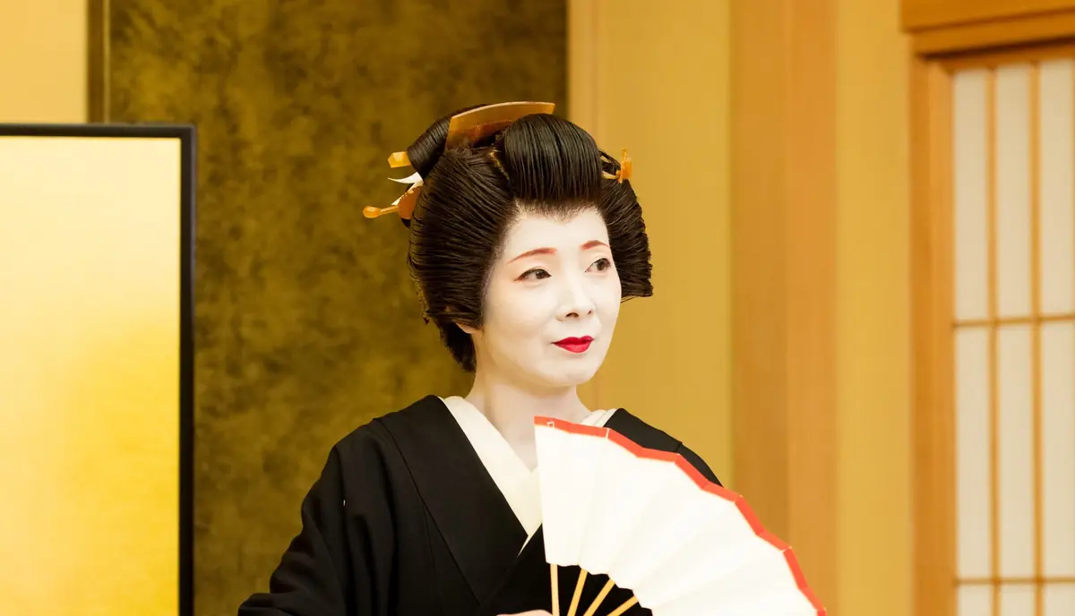 Geisha in a black kimono with white makeup and an ornate hairstyle, holding a folding fan indoors.