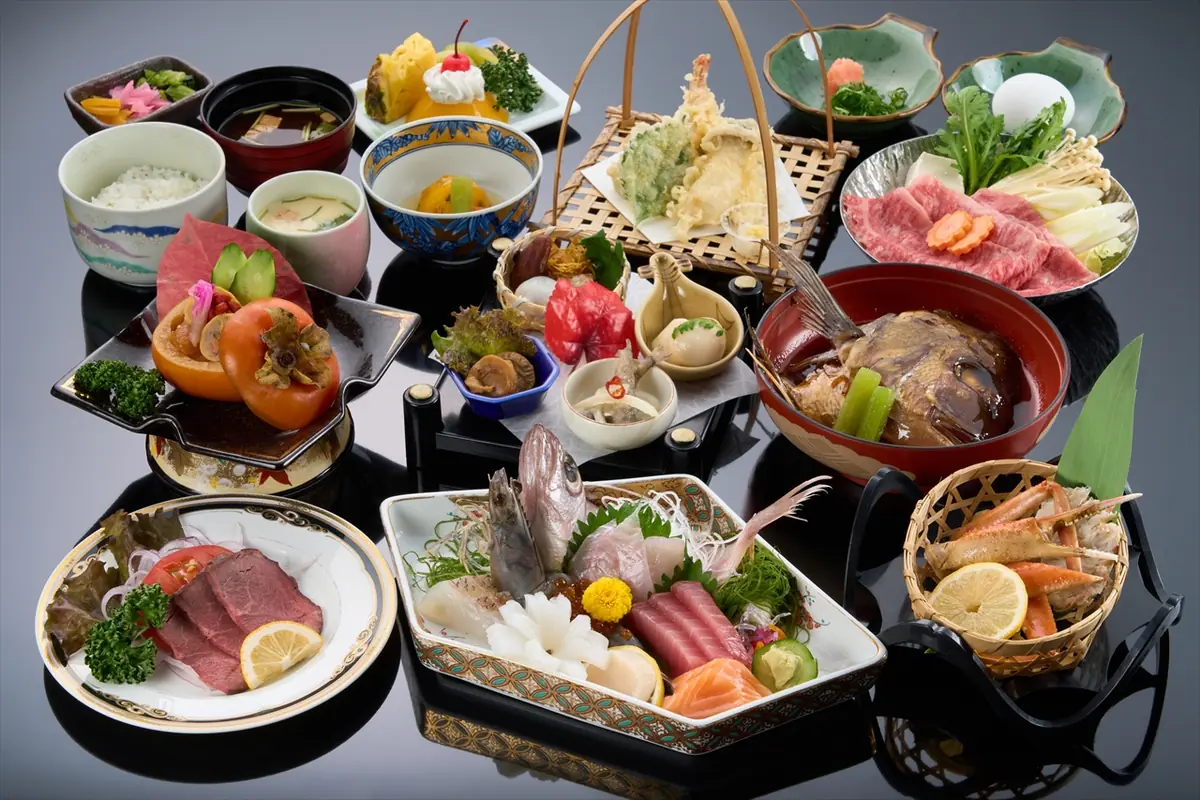 Large spread of Japanese kaiseki: sashimi platter, tempura basket, grilled meat plate, bowls of rice and soup, and assorted small appetizers displayed on lacquerware.