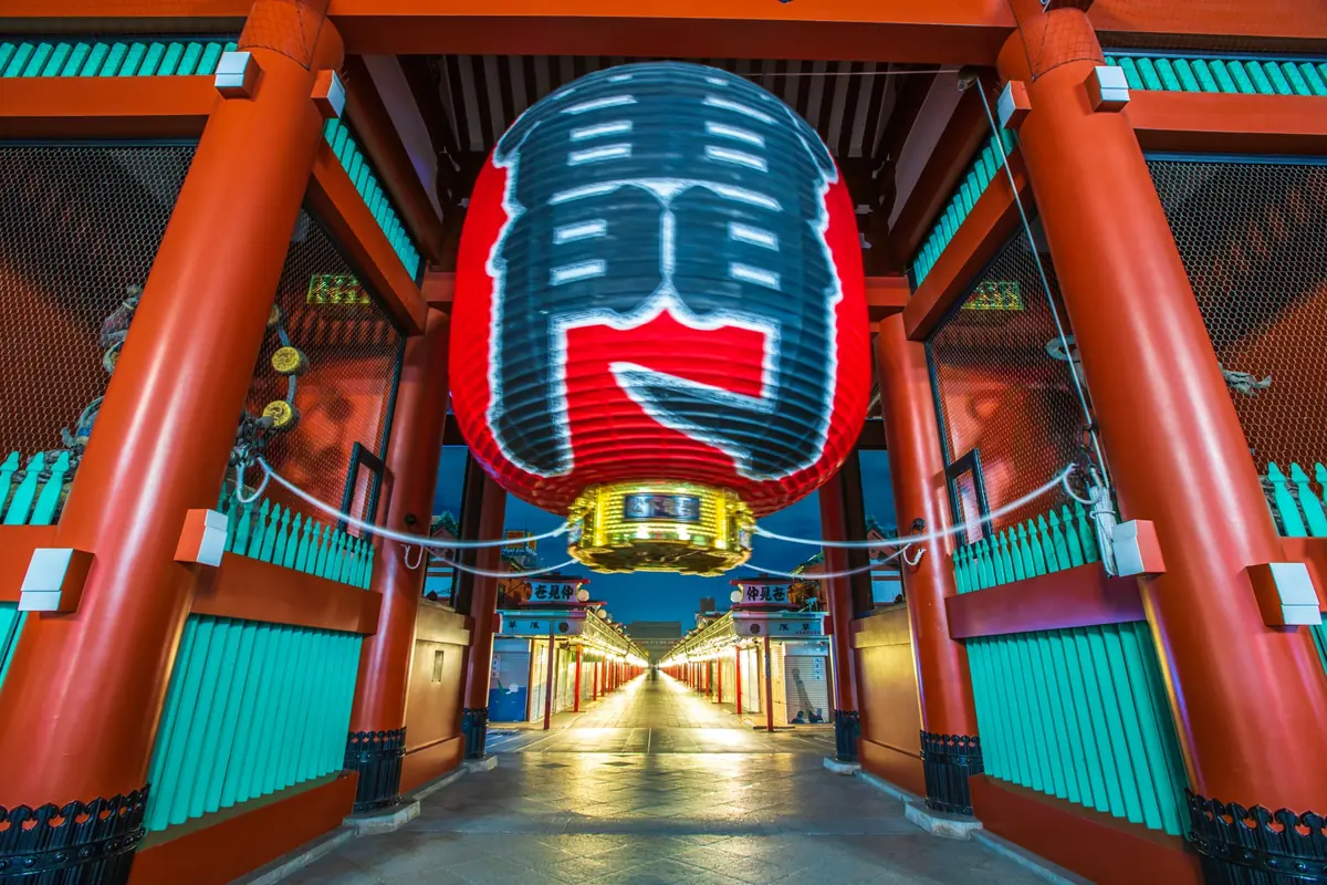 A large red lantern with blue-and-white patterns hangs over a temple gate, framing a bright walkway lined with red pillars and teal fencing.