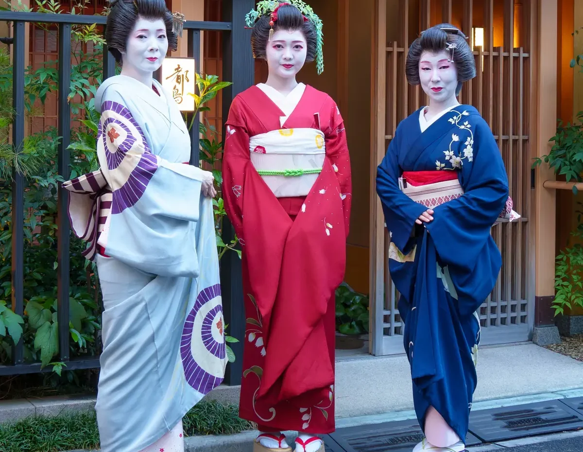 Three women dressed as geisha in colorful kimonos pose outdoors beside a wooden building and greenery.