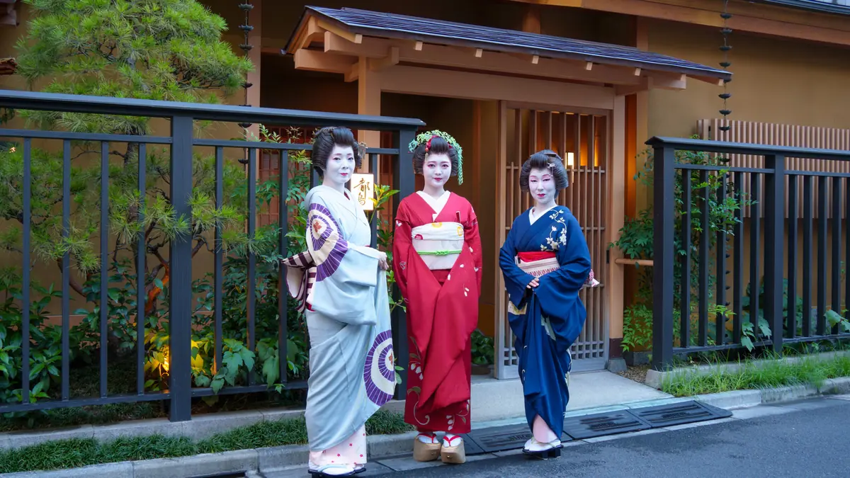 Three geisha in red, blue, and pale gray kimonos pose outside a traditional wooden house with a garden backdrop.