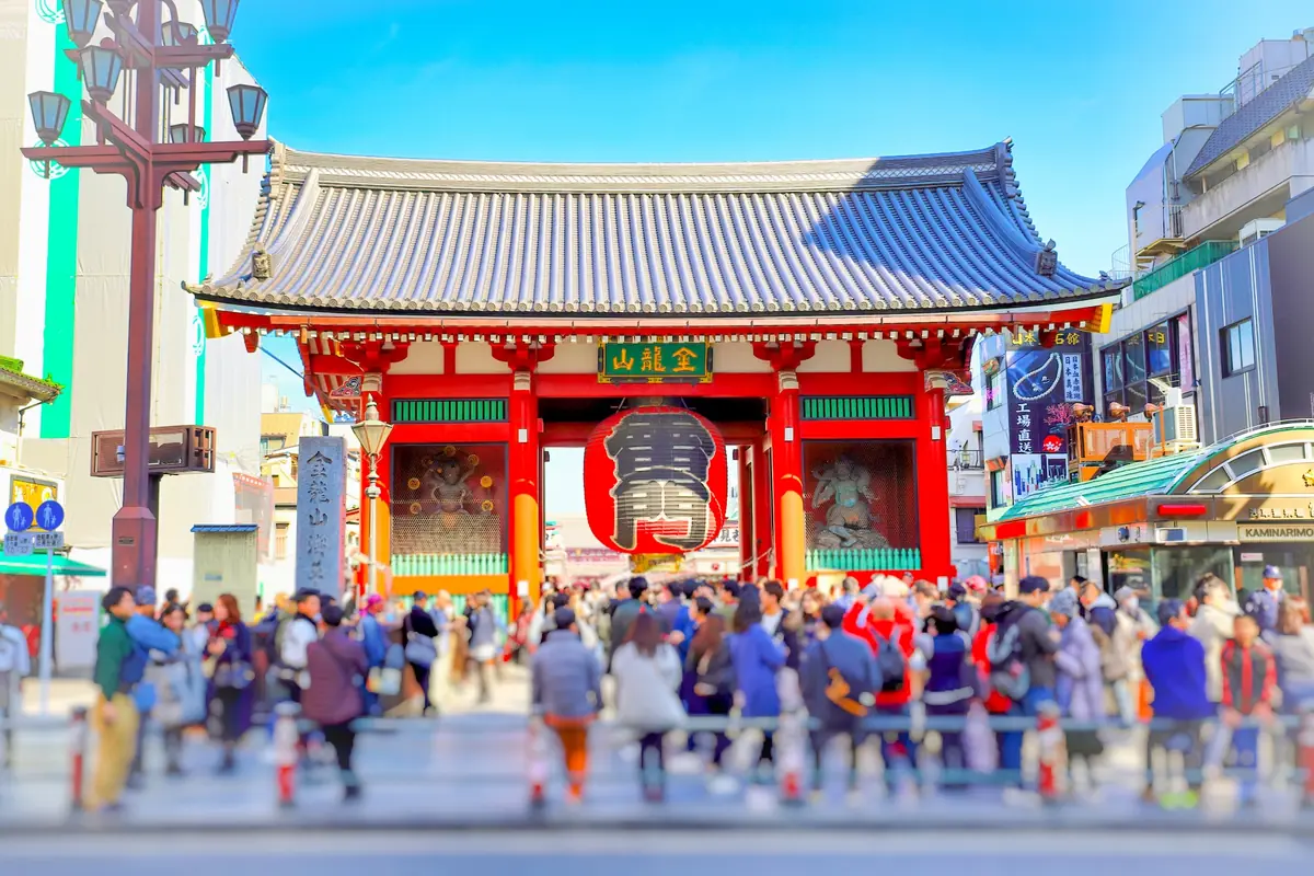 Red temple gate with large hanging lantern at a busy street entrance in Tokyo's Asakusa district, surrounded by pedestrians.