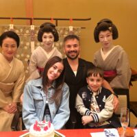 Six people posing for a photo at a celebration: three geisha in traditional kimono behind two smiling visitors, with a birthday cake on the table in front.