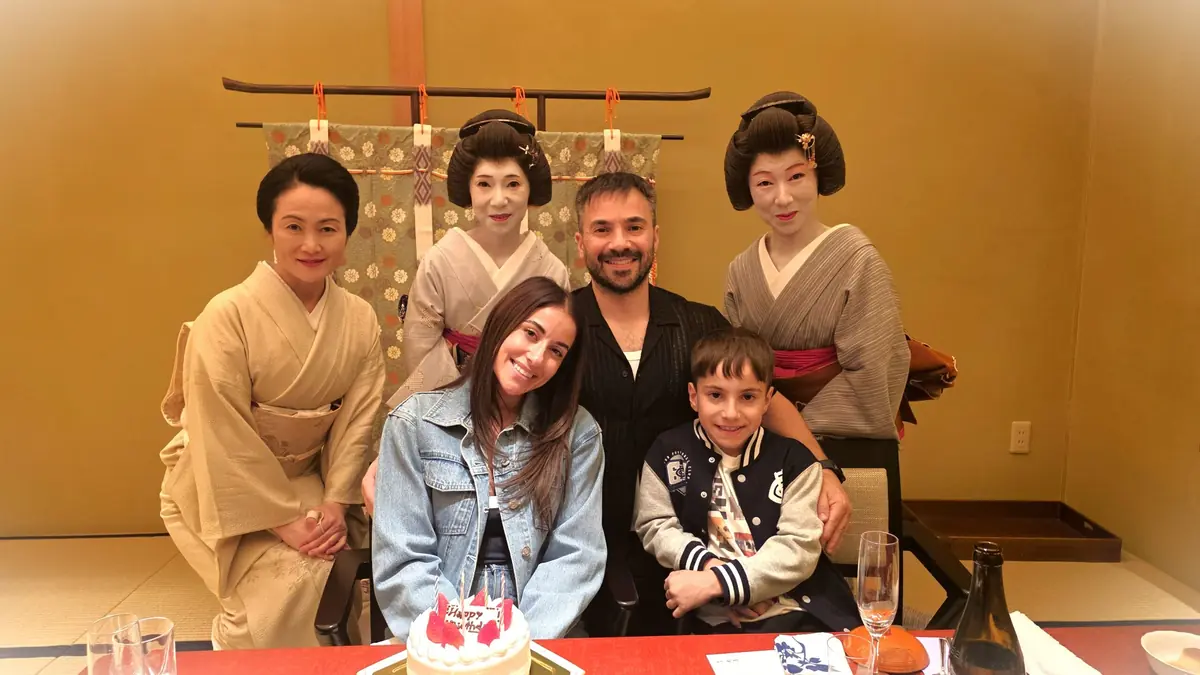 Six people pose for a group photo around a table with a cake: three geisha in kimono stand behind a smiling man, a woman in a denim jacket, and a boy seated in front.