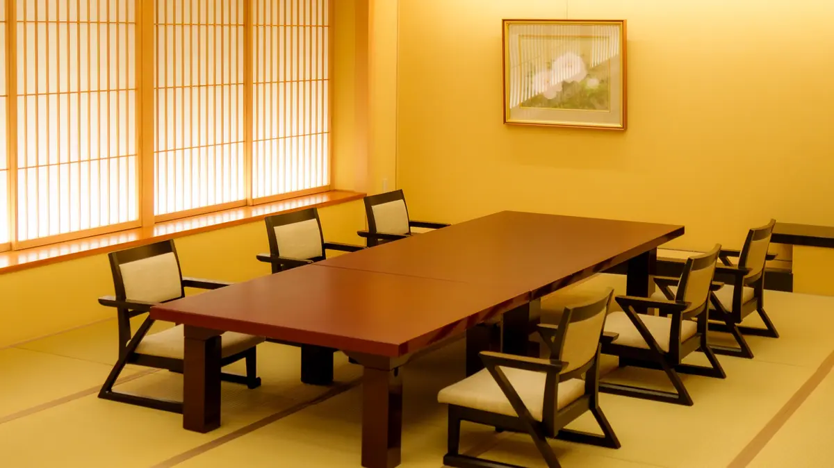 Japanese-style conference room with a long wooden table and beige cushioned chairs, shoji screens along the left wall under warm lighting.