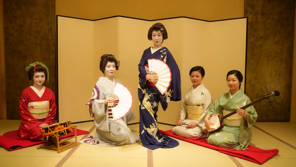 Five women in colorful kimonos posing in a Japanese tatami room with a folding screen; center woman stands with a fan, others sit with fans or instruments (shamisen on the right, small drum on the left).