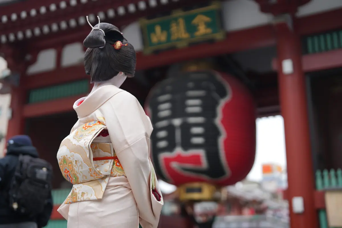Woman in a pale cream kimono with an ornate obi, seen from behind near a large red lantern at a temple entrance.
