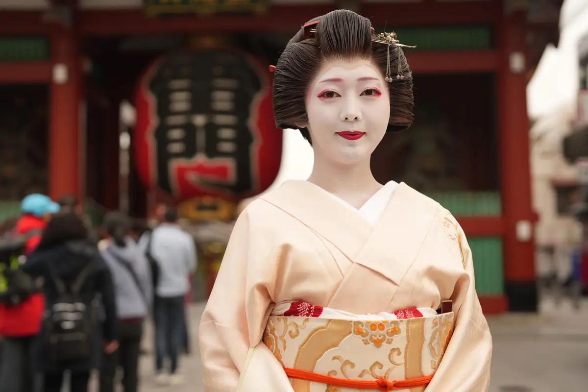 Geisha wearing a pale pink kimono with an ornate obi, standing in a temple courtyard.