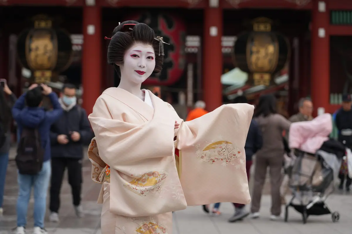 Geisha-style performer with white makeup and elaborate hair in a pale pink kimono, posing in a busy temple courtyard.