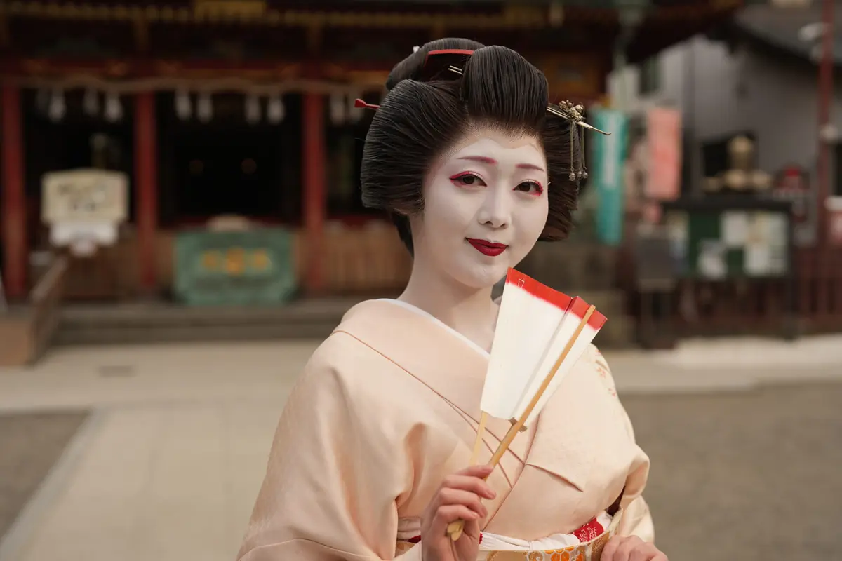 Geisha with white makeup and traditional kimono holding white folding fans outdoors in a temple courtyard