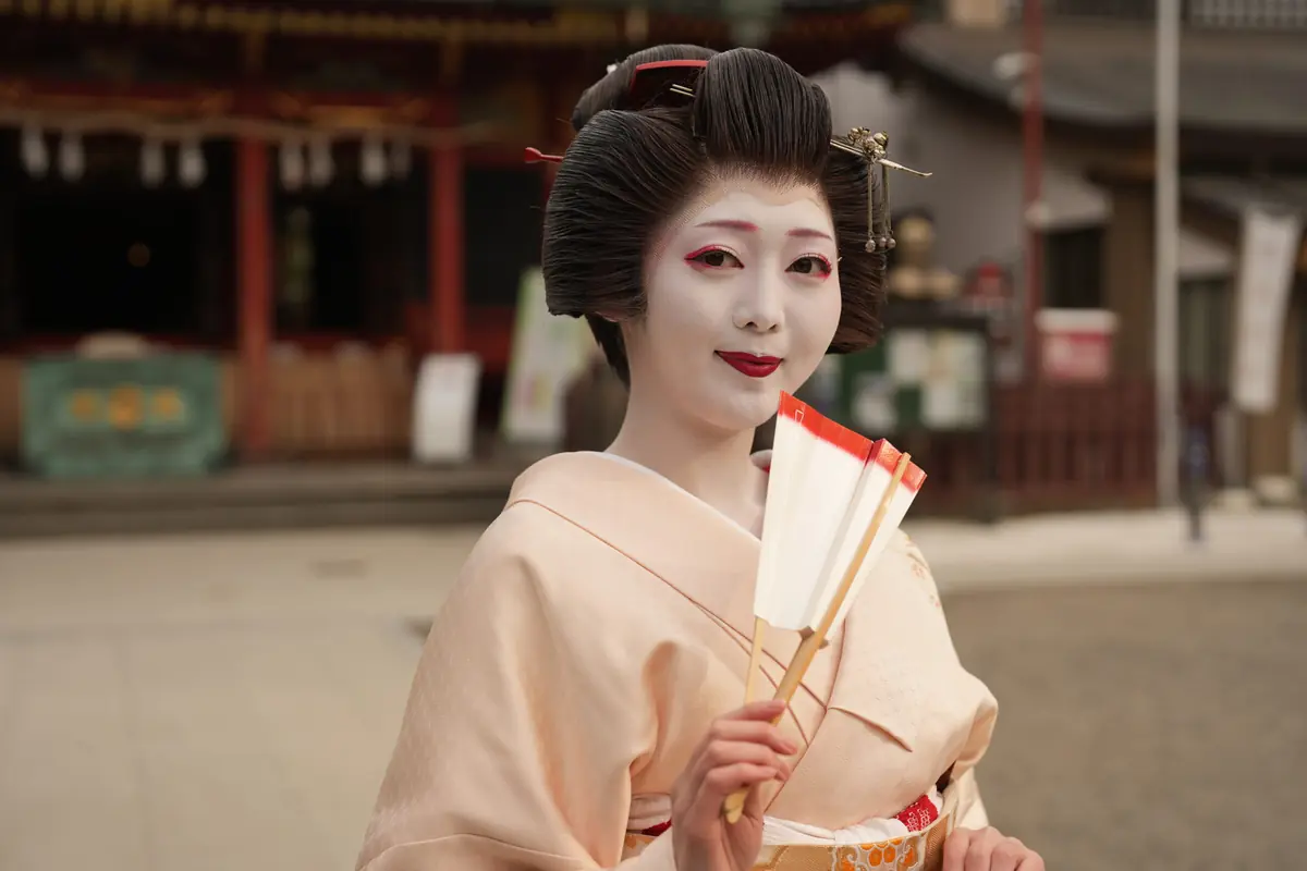 Geisha with white makeup and red lips in a pale pink kimono, holding a folded fan outdoors.
