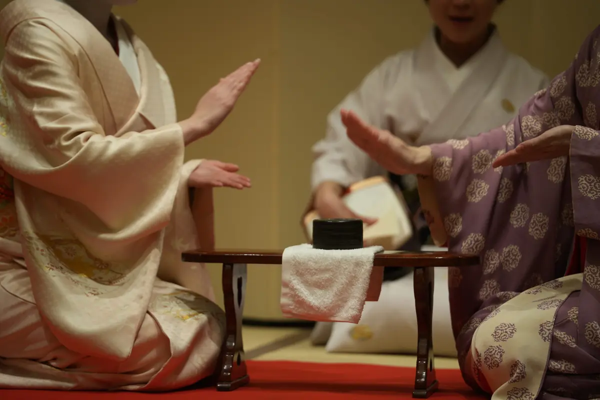 People in traditional robes sit on tatami around a low wooden table, performing a tea ceremony with a black container on a white towel in the center