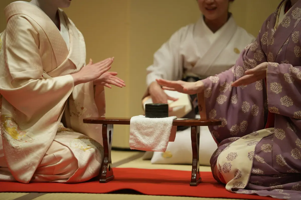 Two people in traditional kimonos kneel at a small wooden table performing a Japanese tea ceremony on a tatami mat with a black tea bowl and towel centerpiece.