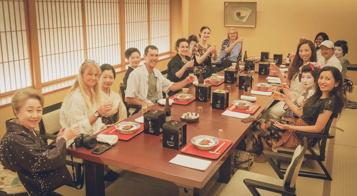 Diverse group of people around a long dining table, toasting with drinks in a Japanese-style room.