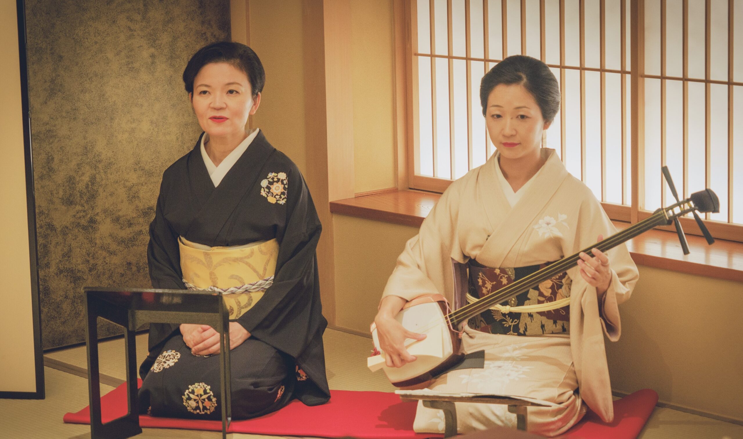 Two geisha musicians (jikata) in an ozashiki — one playing the shamisen while the other performs naga-uta vocals, seated in a tatami room with shoji screens