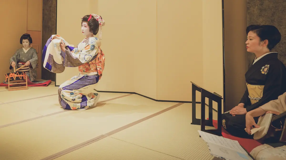 Geisha in a colorful kimono performs a traditional dance on a tatami stage while a musician sits to the left and a spectator watches from the right.