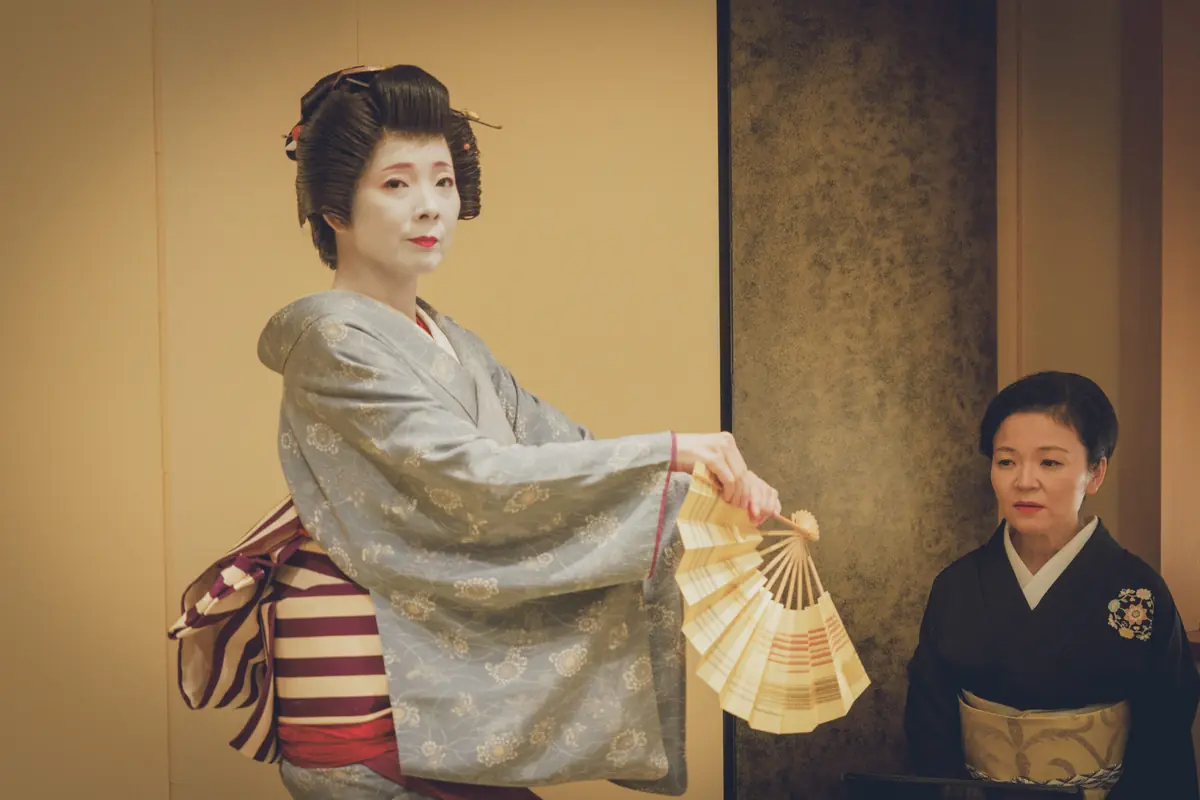 Geisha in a pale blue kimono with white makeup holds a folding fan; an attendant in black watches.