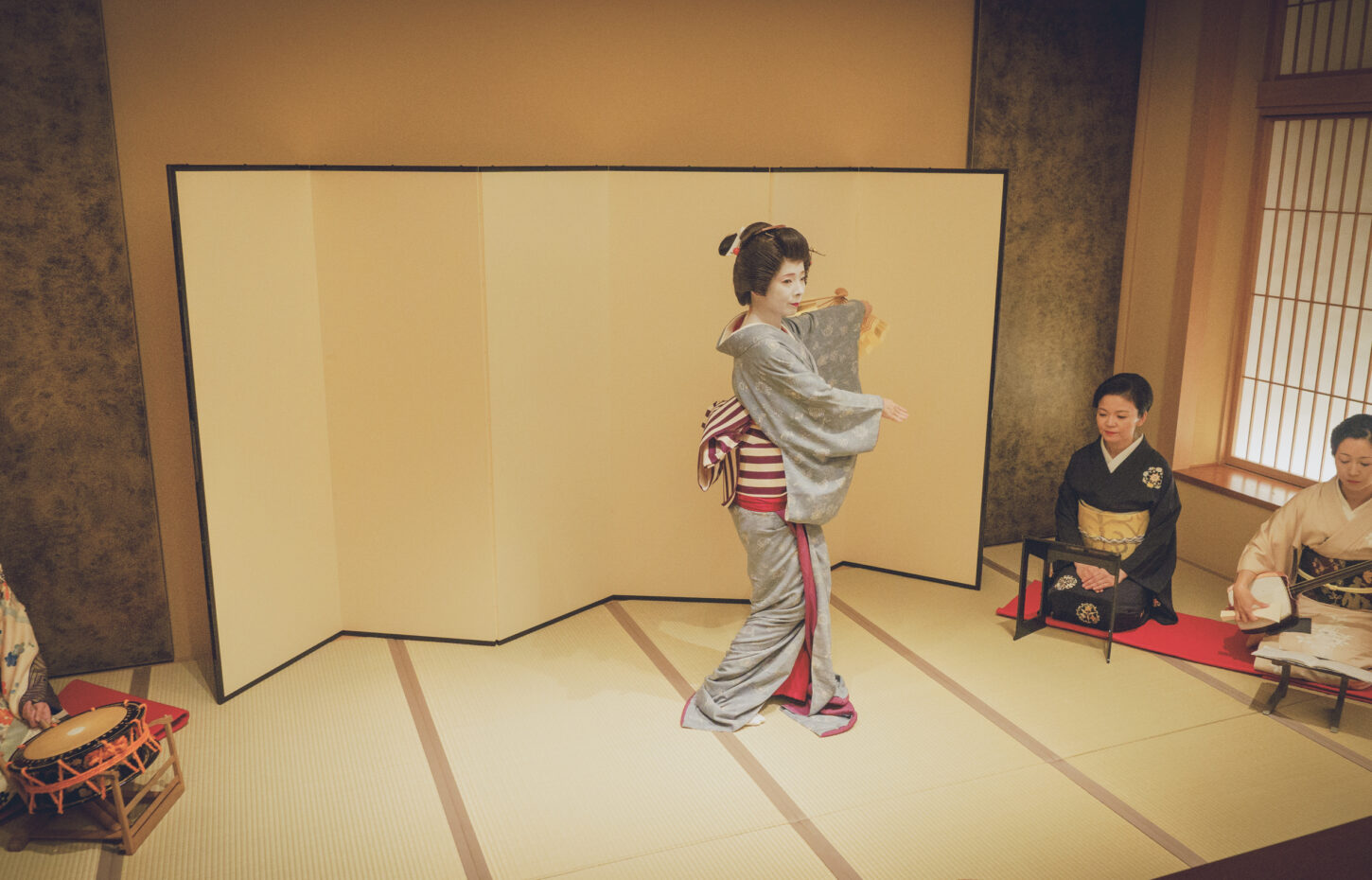Geisha performs a dance in a tatami room as attendants play traditional instruments nearby.