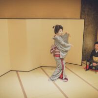 Geisha performs a dance in a tatami room as attendants play traditional instruments nearby.