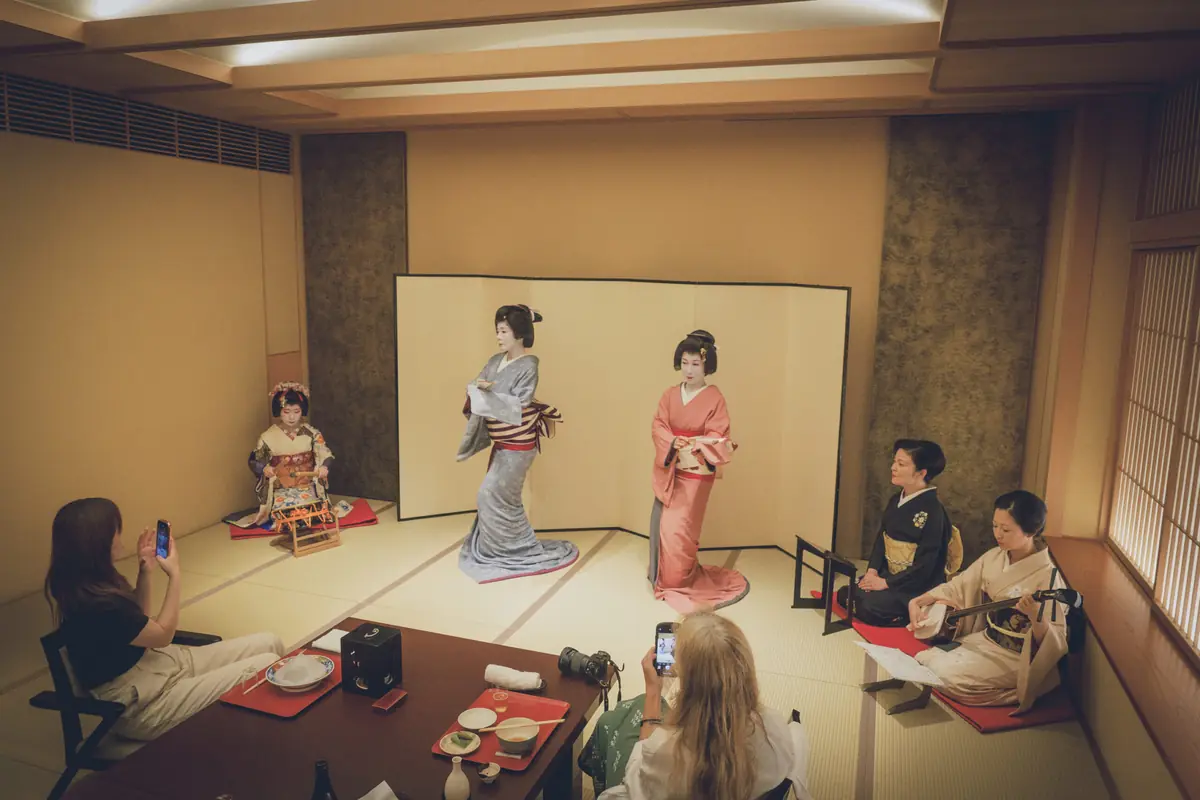 Geisha performers in kimono posing for photos while two musicians sit nearby in a tatami room, audience taking pictures.