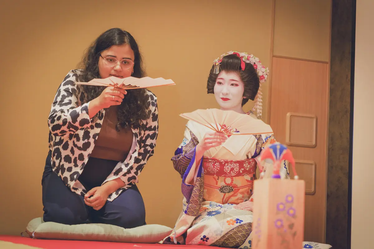 Two women sit side by side indoors; a casually dressed woman with a leopard-print cardigan holds a paper fan, while a geisha in a traditional kimono displays a small fan and makeup.