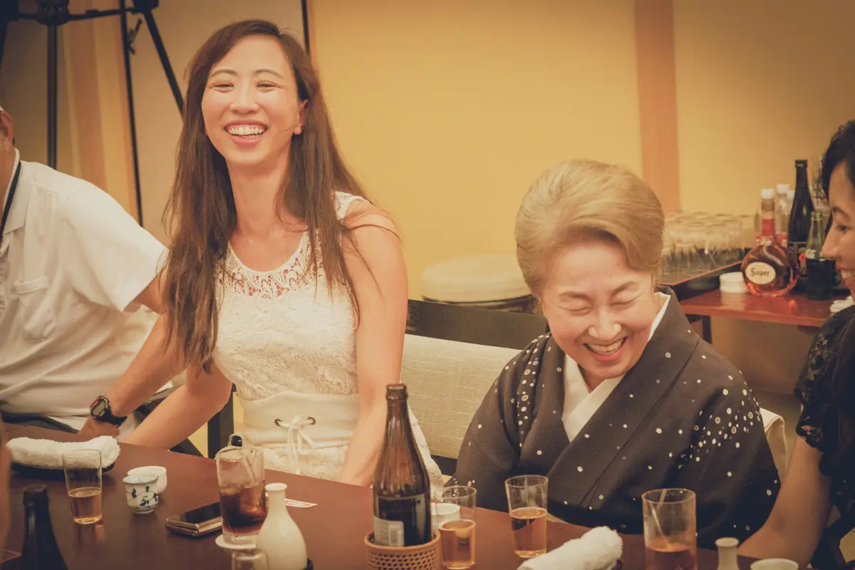 Two women sit at a table, smiling and laughing during a social gathering with drinks and sake bottles nearby.