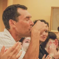 Man in striped shirt kissing his hand in celebration as a group applauds nearby in a warm, indoor setting and yellow walls behind them.