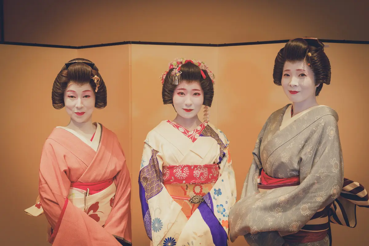 Three geisha in kimono standing side by side against a gold backdrop with elaborate hairstyles.