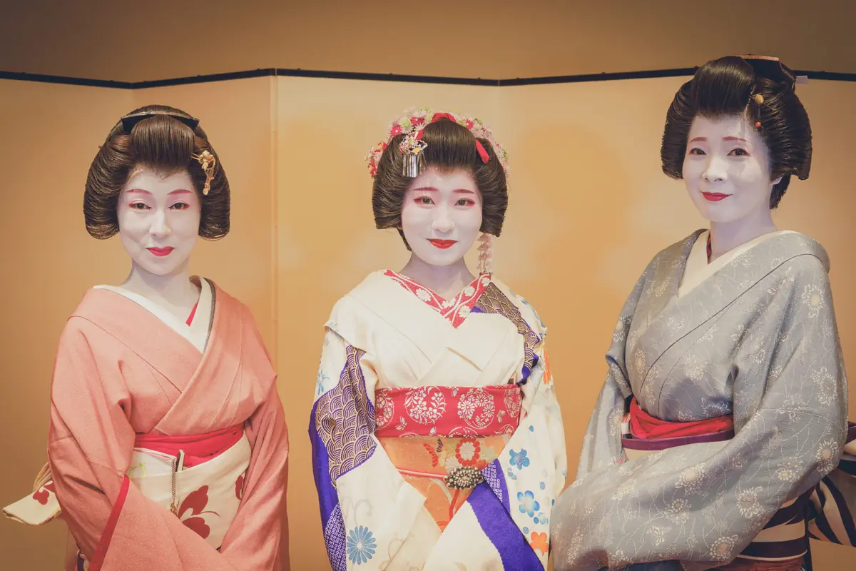 Three women in elaborate kimonos with white makeup and traditional hairstyles pose together for a portrait against a warm gold backdrop.